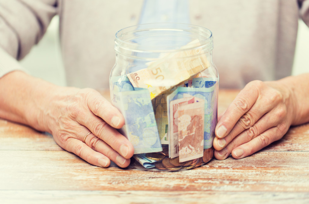 close up of senior woman hands with money in glass jar